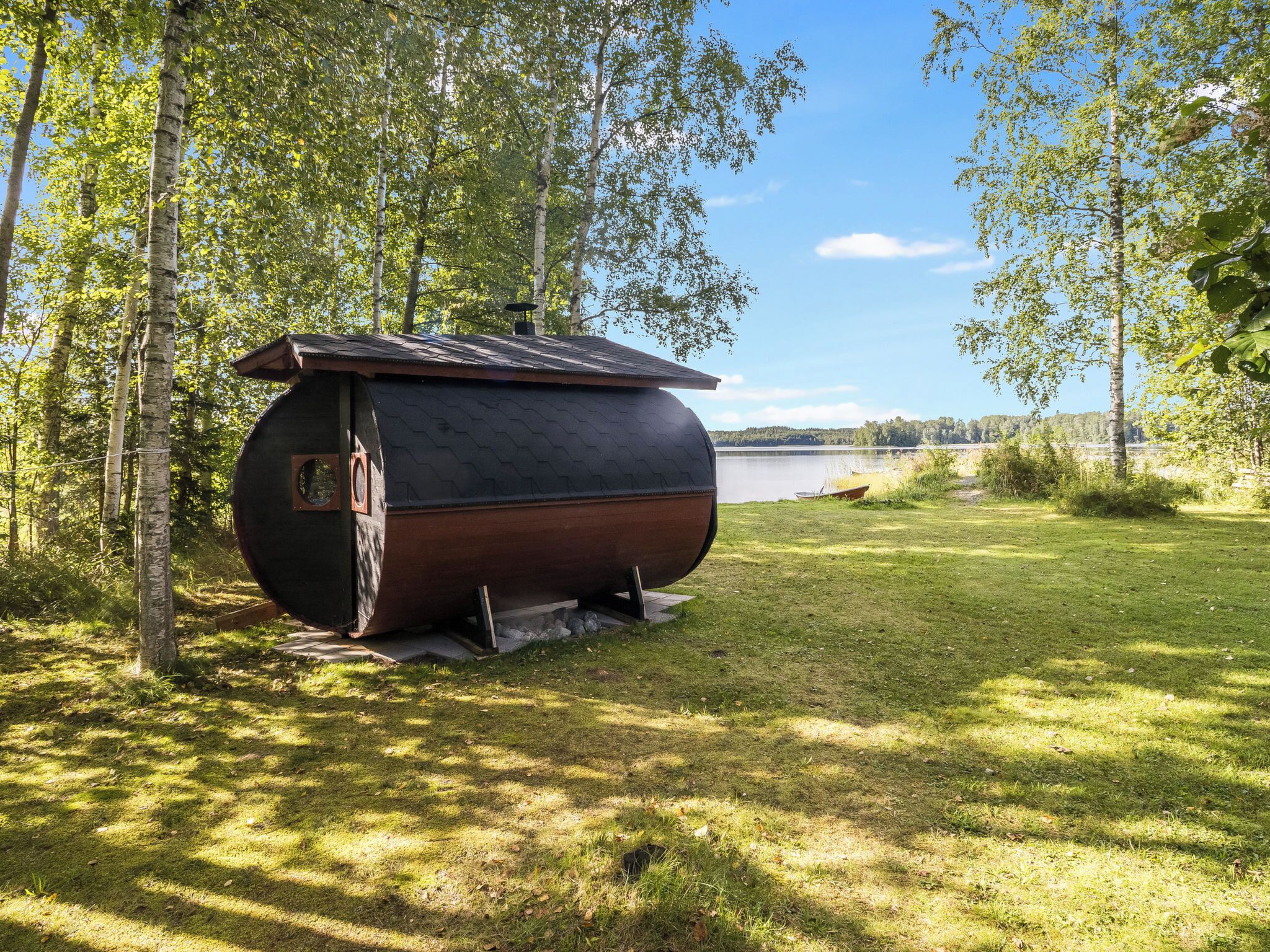 Une grande structure cylindrique surmontée d’un toit en pente, située dans une zone herbeuse, avec des arbres et une étendue d’eau en arrière-plan.