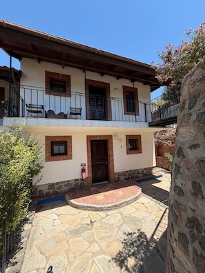 A two-story building with a stone courtyard, a wooden door, and a balcony with a railing.