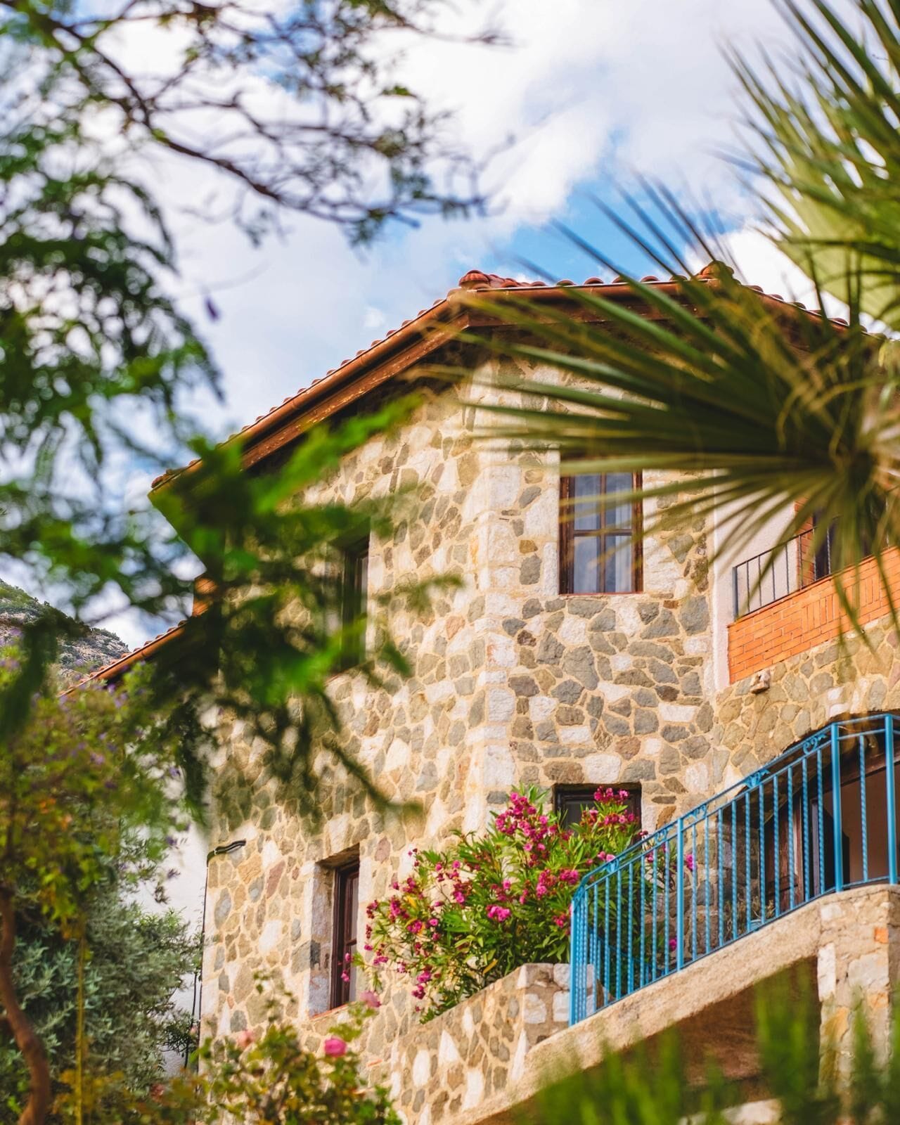 Un edificio de piedra con techo de tejas, un balcón con barandillas azules y flores rosadas en primer plano.