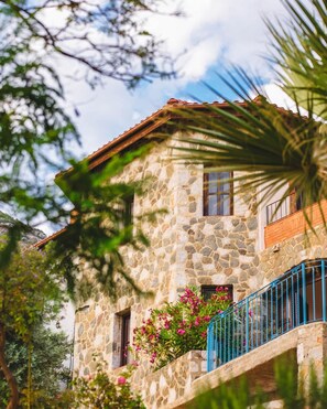A stone building with a tiled roof, a balcony with blue railings, and pink flowers in the foreground.