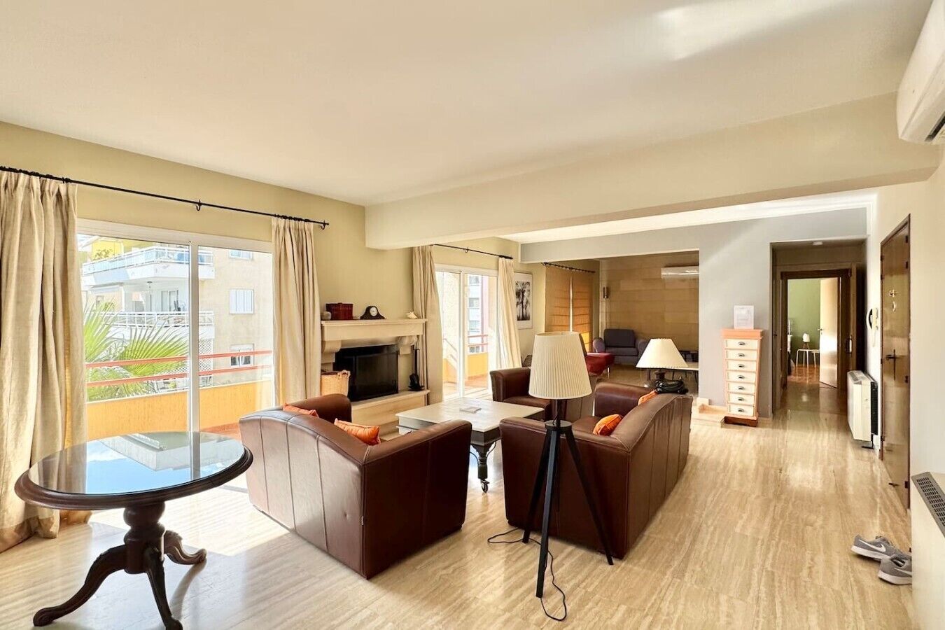 A modern living room with a sofa set, a coffee table, and a TV. There is a dining area with a table and chairs. The room has large windows with curtains.