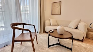 Uma sala de estar moderna com uma cadeira de madeira, uma mesa de centro redonda com um vaso, um sofá bege e uma obra de arte emoldurada na parede.