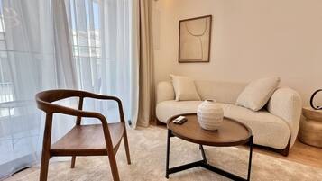 A modern living room with a wooden chair, a round coffee table with a vase, a beige sofa, and a framed artwork on the wall.