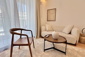 Un salon moderne avec une chaise en bois, une table basse ronde ornée d’un vase, un canapé beige et une œuvre d’art encadrée accrochée au mur.