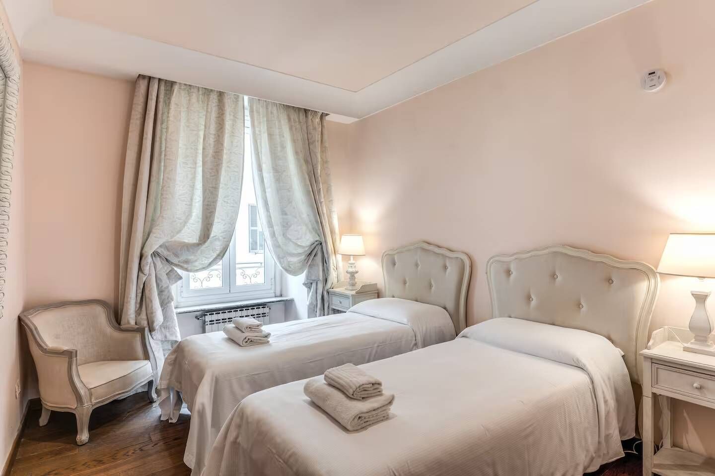 A hotel room with two beds, a chair, a window with curtains, and a radiator.