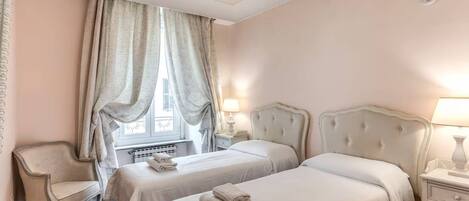A hotel room with two beds, a chair, a window with curtains, and a radiator.