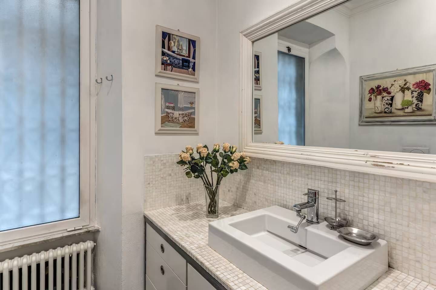 Un baño con un lavamanos blanco, un espejo y un florero con flores. Hay cuadros enmarcados en la pared.