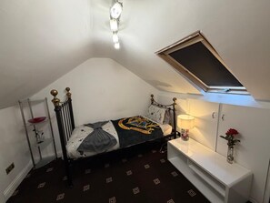 A bedroom with a sloped ceiling, a bed with a headboard, a bedside table with a lamp, and a small shelf with a vase. - Triple Room (Shared House), Near Studios & Station (Watford)