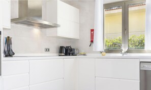 A modern kitchen with white cabinets, a stainless steel range hood, a kettle, a toaster, and a window with a view of greenery. - Villa Albufera by Interhome (Platja de Muro)