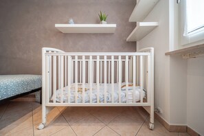 A white crib with a patterned mattress, a small bedside table, and a wall-mounted shelf with a plant. - Laura's 3-bedroom home in the heart of Valeggio sul Mincio (Valeggio sul Mincio)