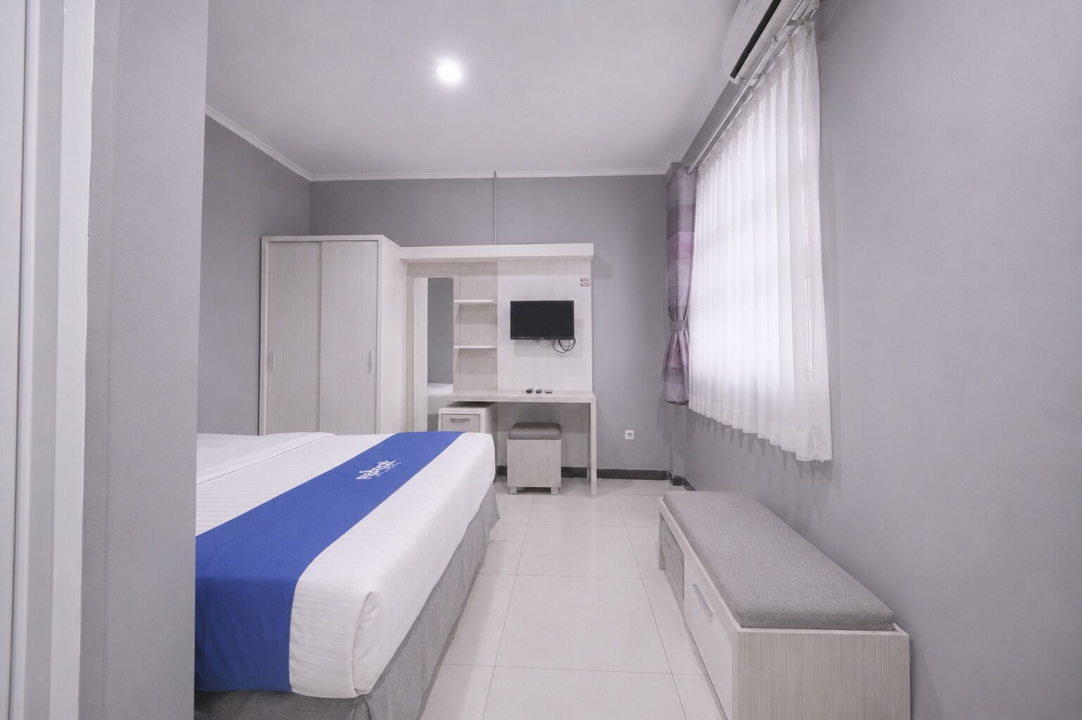 A hotel room with a bed, a desk, a chair, a wardrobe, a television, and a window with curtains.