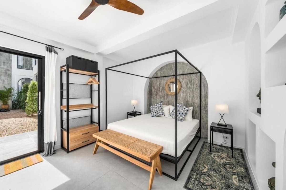 A bedroom with a four-poster bed, a wooden bench, a nightstand with a lamp, and a view of the outdoors.