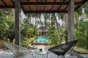 Un área de piscina con sillones de descanso, rodeada de palmeras y una estructura de techo de paja.