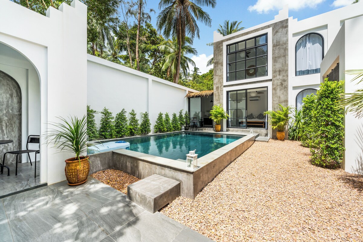 Un área de piscina moderna con deck de concreto, rodeada de vegetación y un edificio blanco con grandes ventanales.