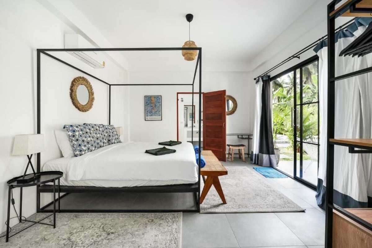 A modern bedroom with a four-poster bed, a bedside table, a bench, and a view of greenery through large windows.