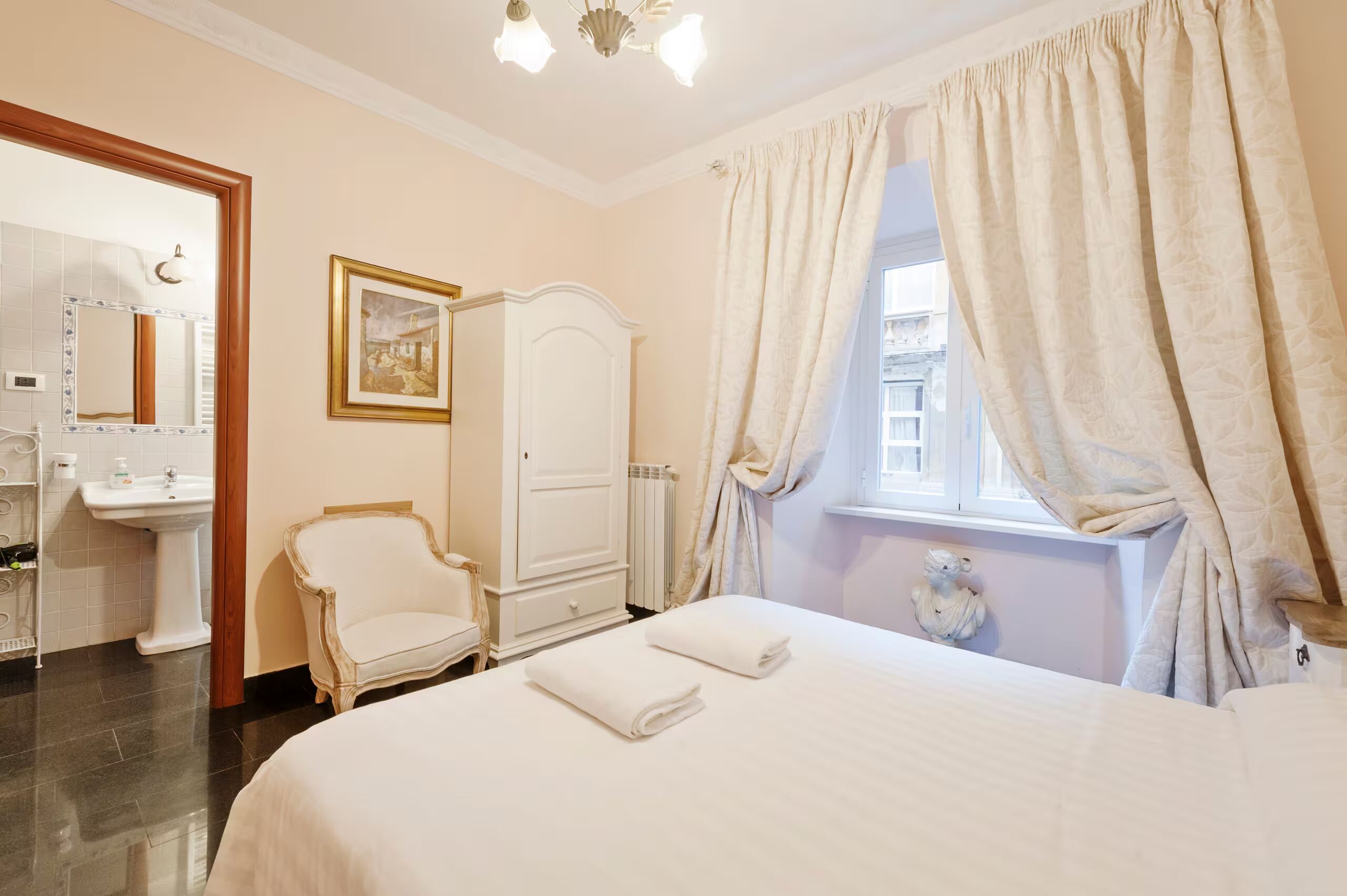 Une chambre d’hôtel comprenant un lit, une chaise, une armoire, une salle de bain avec un miroir et un lavabo, et une fenêtre avec des rideaux.