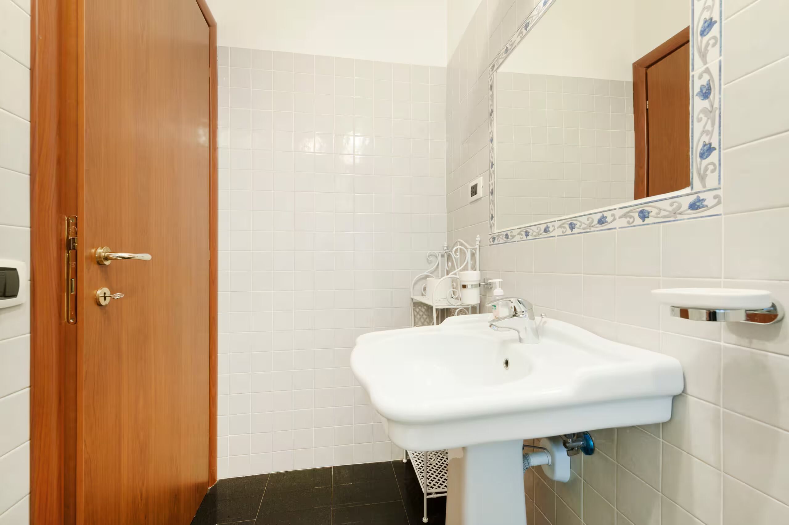 A bathroom with a wooden door, white tiled walls, a white sink, and a mirror with decorative tiles.