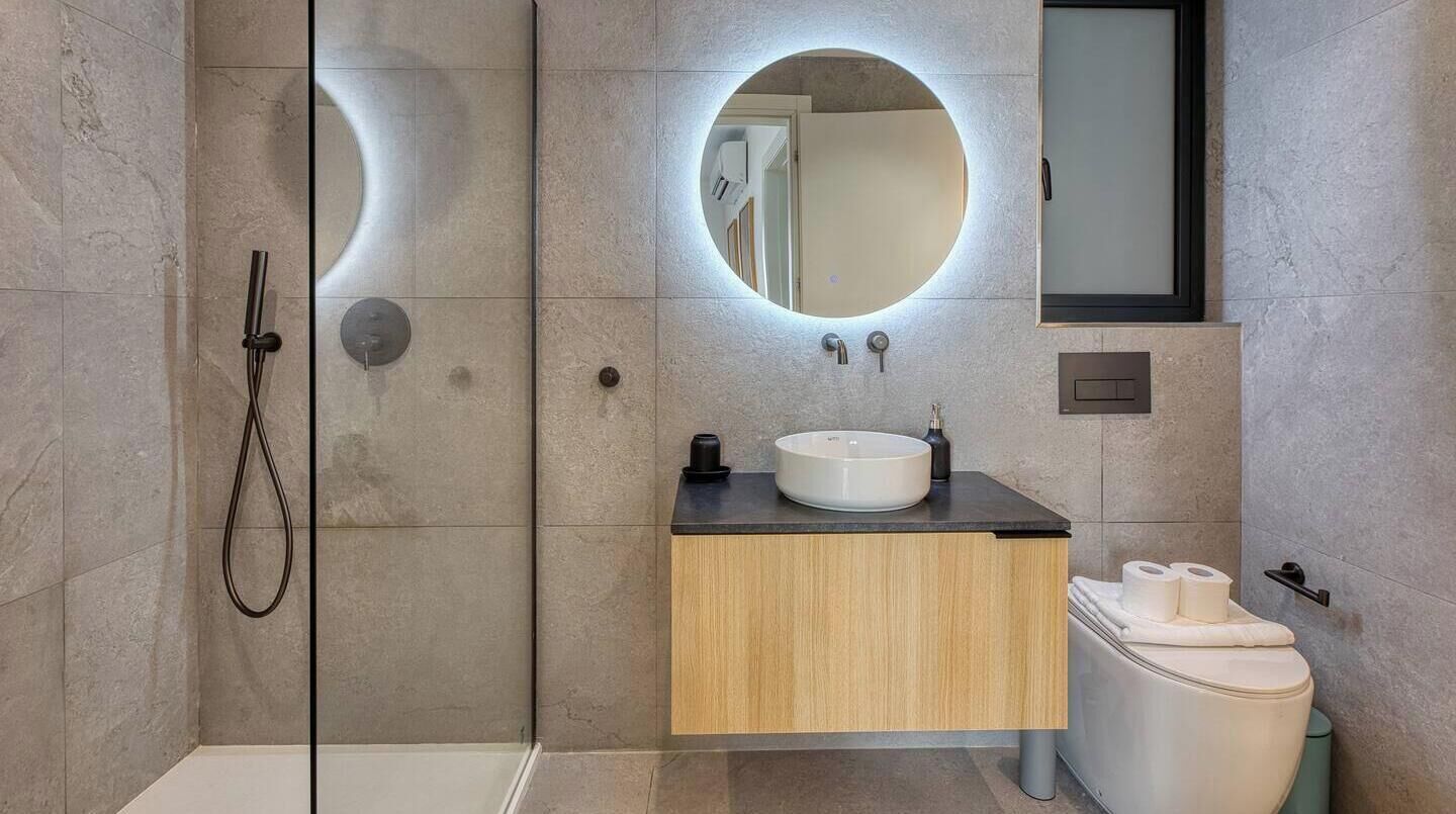 Un bagno moderno con doccia a vetro, mobile bagno in legno con lavandino bianco e WC con portasci asciugamani.