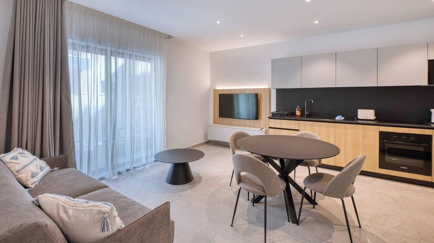 Un soggiorno moderno con un divano, un tavolo da pranzo rotondo e sedie. È presente una zona cucina con mobili e elettrodomestici.