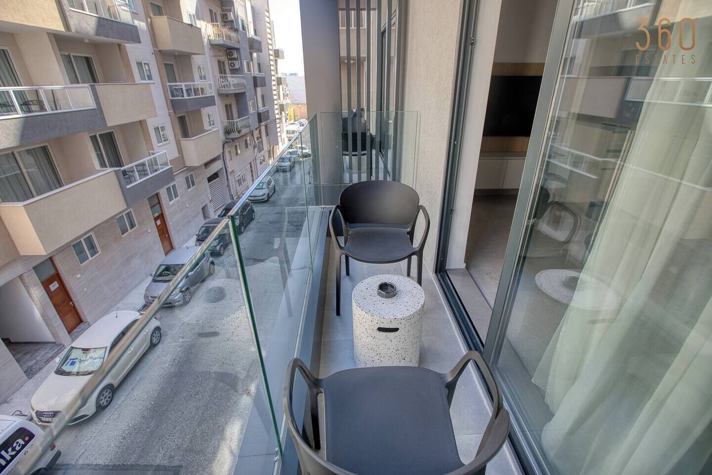 A balcony with two chairs and a small round table, overlooking a street with parked cars and buildings.