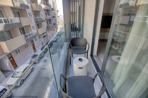 Varanda com duas cadeiras e uma mesa redonda pequena, com vista para uma rua com carros estacionados e edifícios.