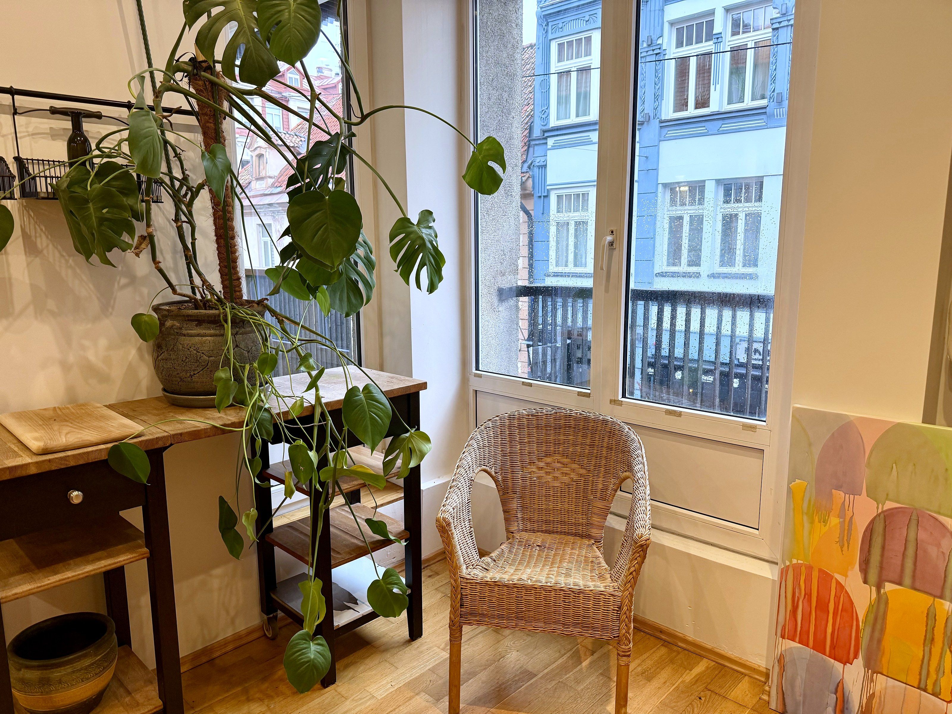 A room with a wooden table, a wicker chair, a potted plant, and a window with a view of buildings.