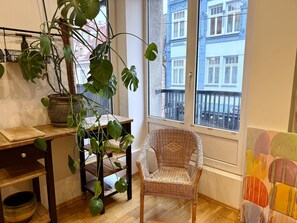 A room with a wooden table, a wicker chair, a potted plant, and a window with a view of buildings.