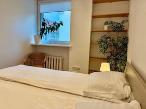A bedroom with a bed, a window, a radiator, a chair, and shelves with plants. - Pilies23 Old Town Luxury Apartment (Vilnius)