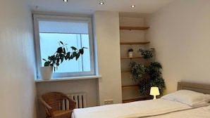 A bedroom with a bed, a chair, a window with plants, and built-in shelving.