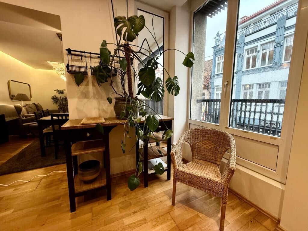 Ein Zimmer mit Esstisch, Stühlen, einem Korbsessel, einer Hängepflanze und Blick auf ein Gebäude durch ein Fenster.