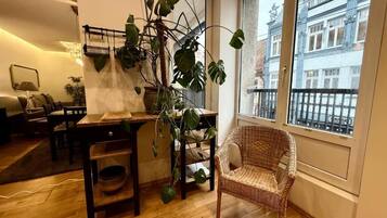 Ein Zimmer mit Esstisch, Stühlen, einem Korbsessel, einer Hängepflanze und Blick auf ein Gebäude durch ein Fenster.