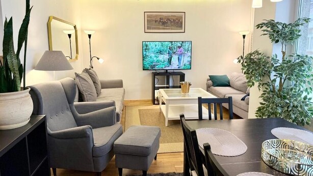 A modern living room with a grey sofa, a coffee table, and a TV. There are plants and a chandelier.
