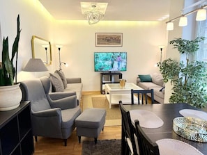 A modern living room with a grey sofa, a coffee table, and a TV. There are plants and a chandelier. - Pilies23 Old Town Luxury Apartment (Vilnius)