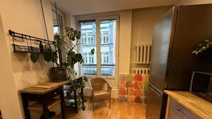 A kitchen with a wooden floor, a refrigerator, a dining table with chairs, and a window with a view of buildings.