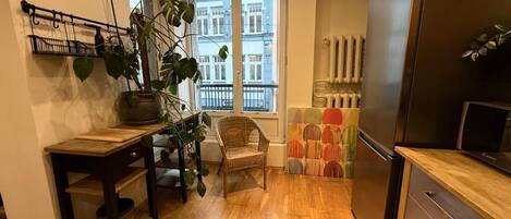 Una cocina con piso de madera, un refrigerador, una mesa de comedor con sillas y un ventanal con vista a edificios.