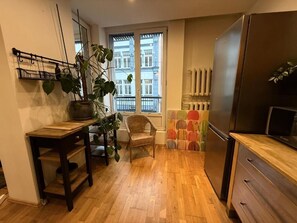 Una cocina con piso de madera, un refrigerador, una mesa de comedor con sillas y un ventanal con vista a edificios.