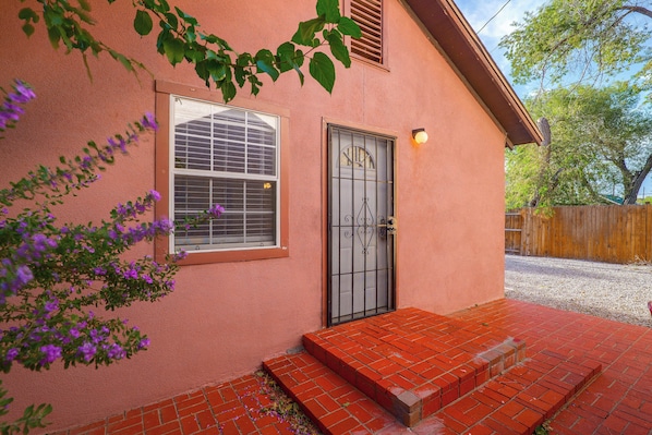 A house with a red brick entrance, a window with blinds, a door with a metal gate, and a wooden fence in the background. - Couple’s Casita Near Historic Downtown Roswell! (Roswell)