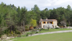 A two-story house with a stone wall, surrounded by a forested area.