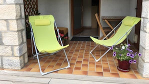 Two green lounge chairs on a tiled patio with a wooden table and a potted plant with purple flowers.