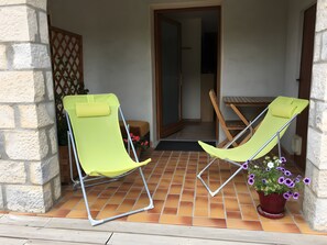 Two green lounge chairs on a tiled patio with a wooden table and a potted plant with purple flowers.