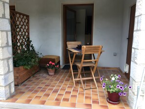 A patio with a wooden table and chair, potted plants, and a tiled floor.
