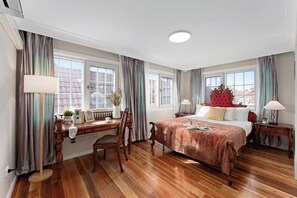 A hotel room with a wooden floor, a bed with a red headboard, a desk with a chair, and two windows with curtains. - Charming Character Terrace, Eclectic Vintage Charm (Petersham)