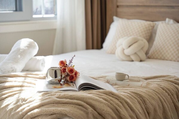 A neatly made bed with a blanket, a vase with flowers, a cup, and an open book.