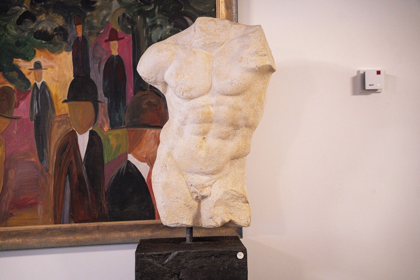 Een sculptuur op een voetstuk in een kunstgalerie, met een schilderij op de achtergrond.