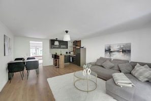 A modern living room with a grey sofa, a dining area with a table and chairs, and a kitchen in the background.