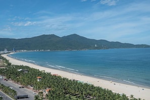 A coastal road with palm trees, a beach, and a mountain range in the background. - Coast & Nest apartment (Đà Nẵng)