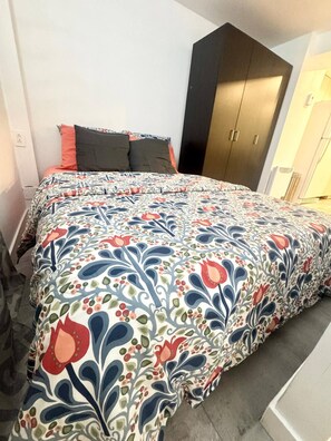 A bed with a floral-patterned bedspread, a dark-colored wardrobe, and a white wall with a power outlet.