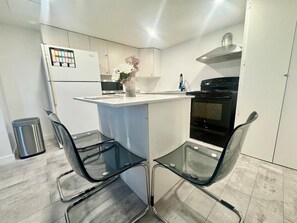 A modern kitchen with a central island, two glass chairs, a stainless steel trash bin, and a black stove.