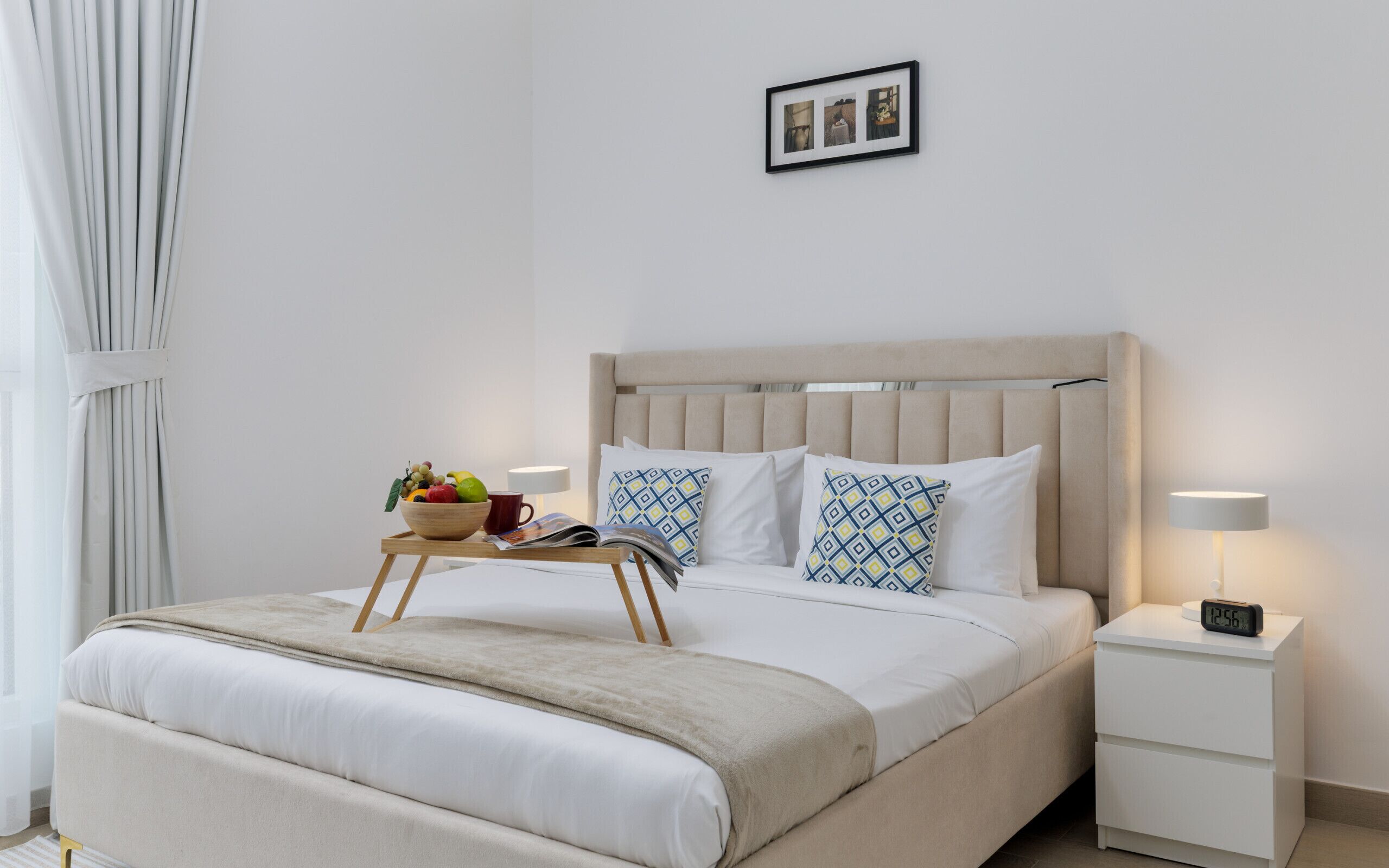 Un lit bien fait, avec une tête de lit beige, une table de chevet sur laquelle se trouve une lampe et une horloge numérique, et un petit plateau en bois contenant des fruits et une tasse rouge.