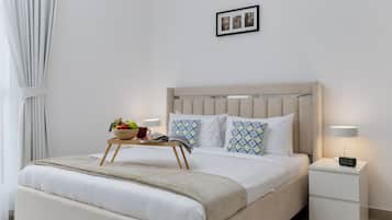 Un lit bien fait, avec une tête de lit beige, une table de chevet sur laquelle se trouve une lampe et une horloge numérique, et un petit plateau en bois contenant des fruits et une tasse rouge.
